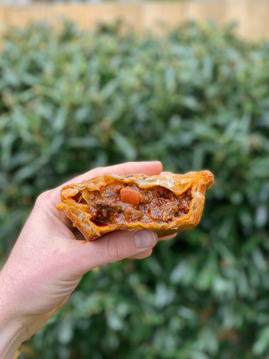 Left hand holding Beef Rendang Roti Pie with bites taken out showing the filling. Green hedge out of focus in the background.